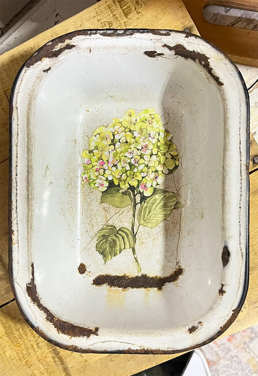 Rectangular white enamelware pan with rust and a black rim repurposed as a decor piece with light green and yellow hydrangea branch motif.