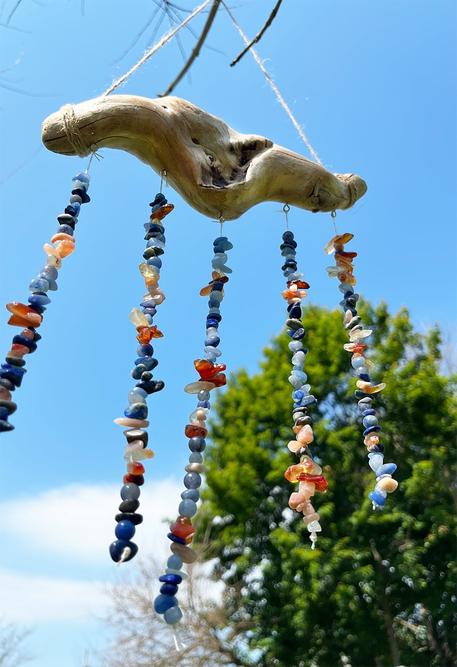 Colorful strands of beads hanging from a weathered piece of driftwood against a blue summer sky, creating a handmade mobile-inspired outdoor decoration.