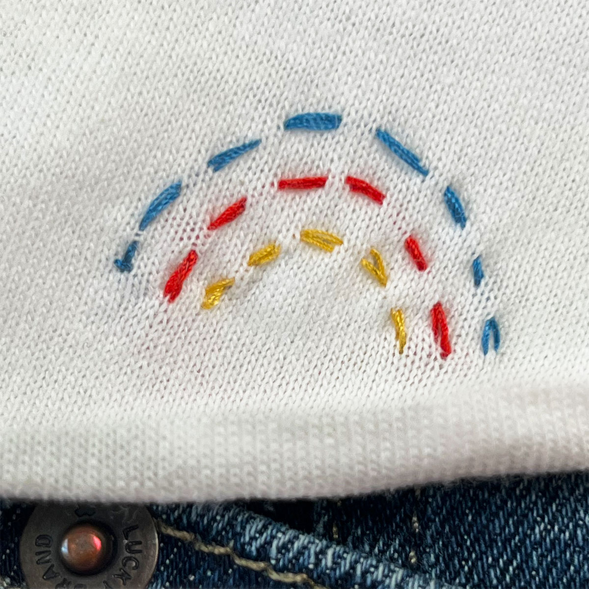 Closeup of a tiny running stitch rainbow in blue, red and yellow thread stitched onto the bottom of a white t-shirt with a hint of denim below.