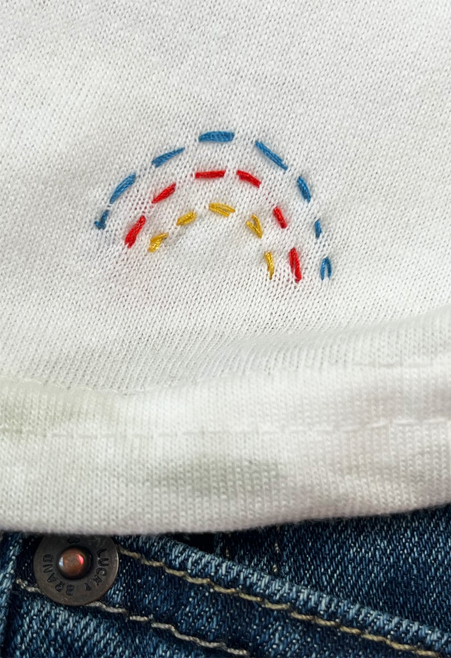 Closeup of a tiny running stitch rainbow in blue, red and yellow thread stitched onto the bottom of a white t-shirt with a hint of denim below.