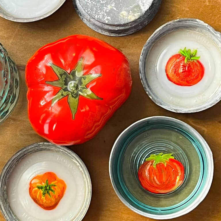 Tomatoes painted on the inside of old glass and metal canning jar lids. In the center is a ceramic tomato red heirloom tomato. One of the tomatoes is painted on a blue glass canning jar lid. Two others on the white glass.
