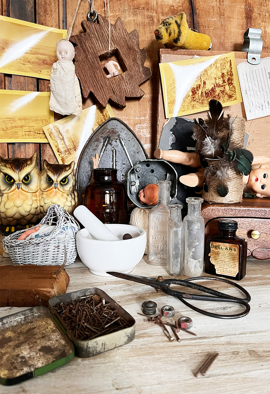 Oddities on work table and wall including a set of judging owls, a wood cog, vintage babies, feathers, stone mortar and pestle, glass jars and vintage elixir bottles, rusty tin of nails, vintage scisors and old yellowed polaroids hanging.