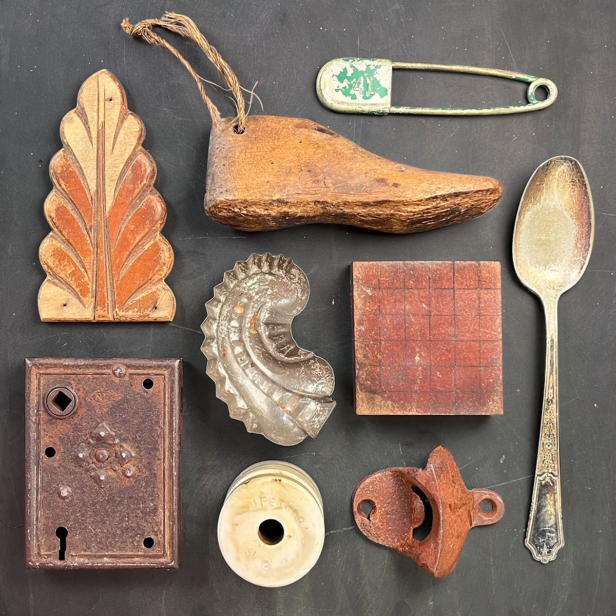 Assorted vintage found objects arranged on a dark slate surface, including a carved wooden ornament filigree, shoe last, large military safety pin, tarnished spoon, metal mold fragment, gridded wood block, antique lock plate, ceramic insulator, and a rusted bottle opener.