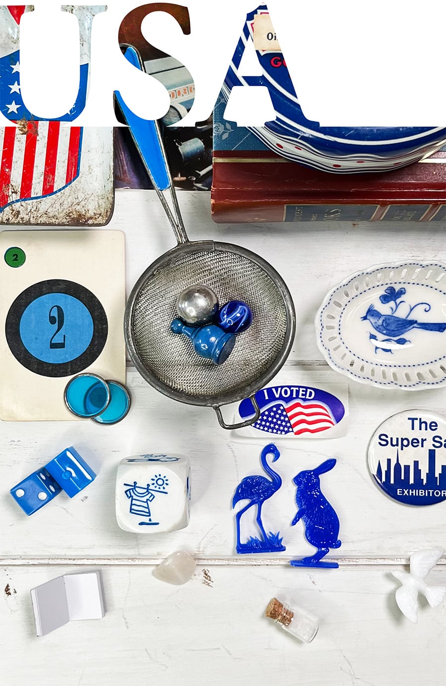 Vintage-inspired Americana flatlay featuring oversized USA typography filled with blue objects, including game pieces, glass marbles, dominoes, mini blue rabbit and flamingo, a bluebird dish, and nostalgic summer details arranged on white shiplap wood.