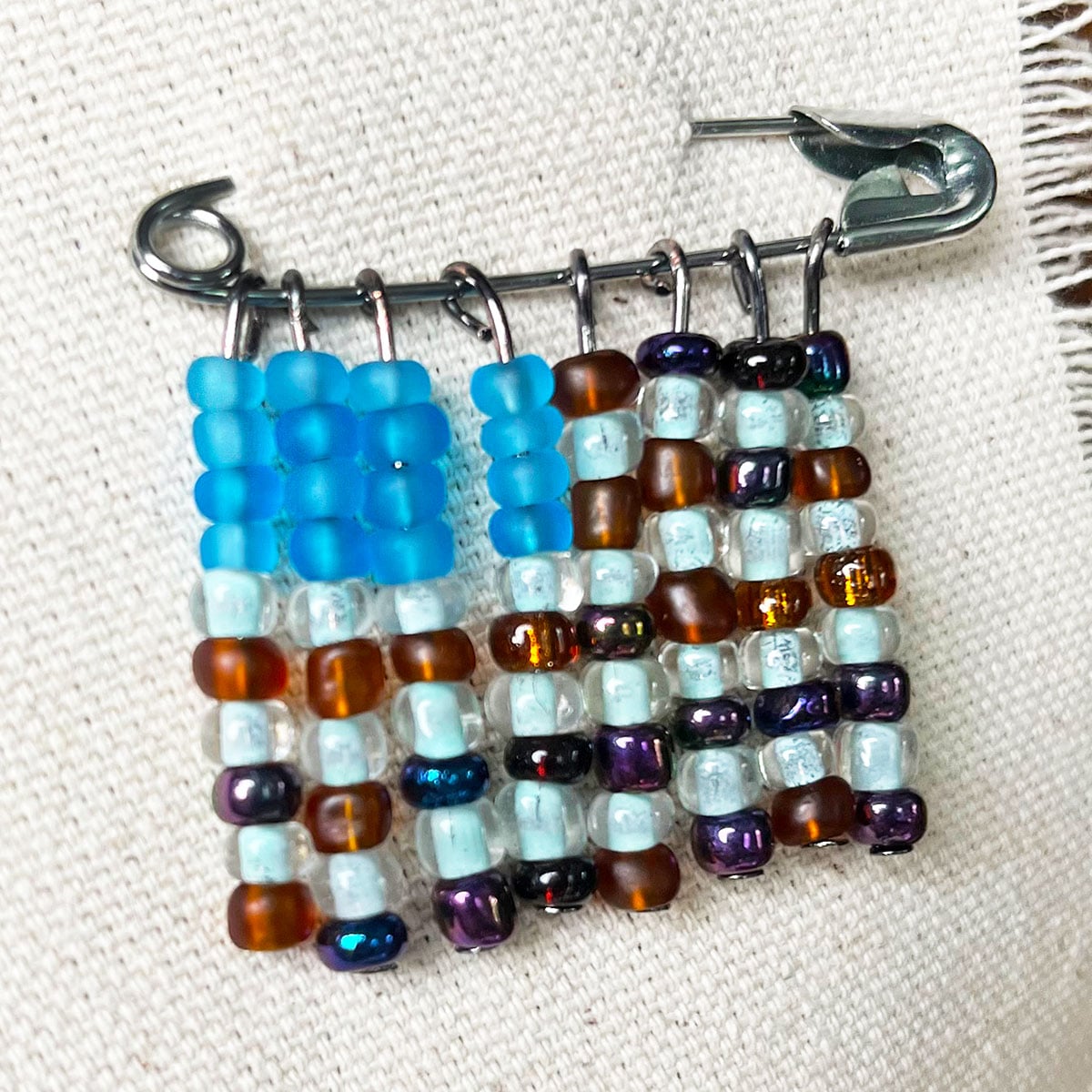 Closeup photo of a handmade seed bead american flag pin made with wire strands to stack the beads hanging from a large silver safety pin. The bead colors are amber for the red stripes and ocean blue for the blue flag portion with transparent pearl for the white stripes.