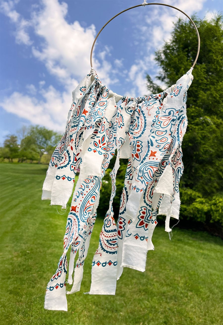 A wire hoop hung outdoors with torn cream bandana strips blowing in the breeze against a blue summer sky and green trees, creating a soft Americana-inspired textile display.