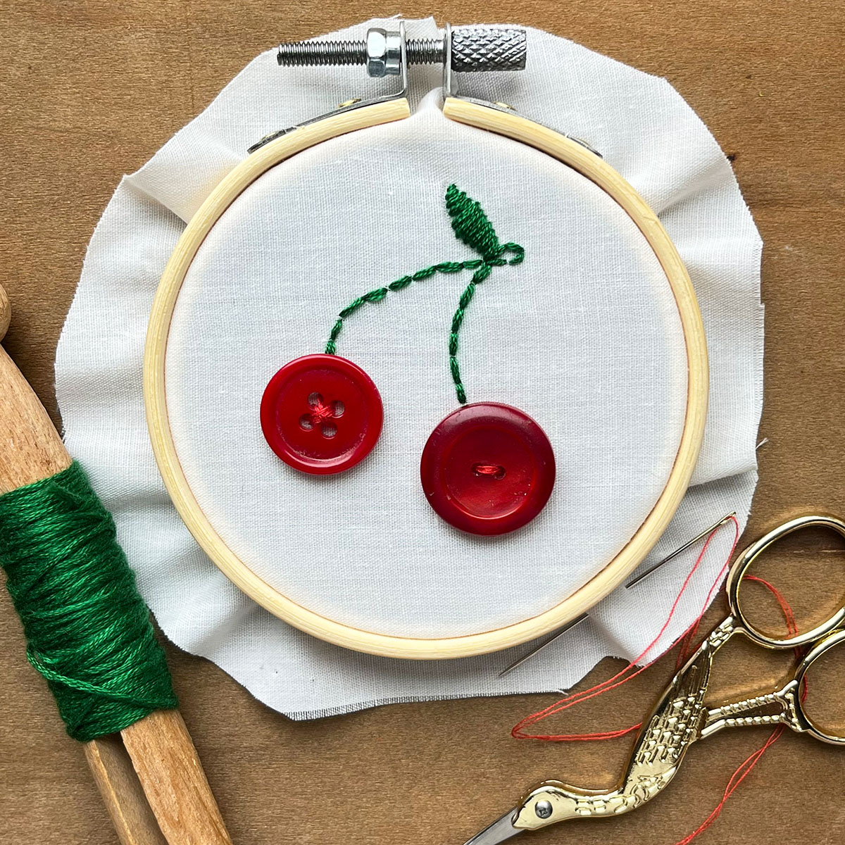 Three inch wooden hoop with ivory fabric embroidery cherry stems in green thread with on leaf at the top and two different red buttons for cherries on a warm wood background.