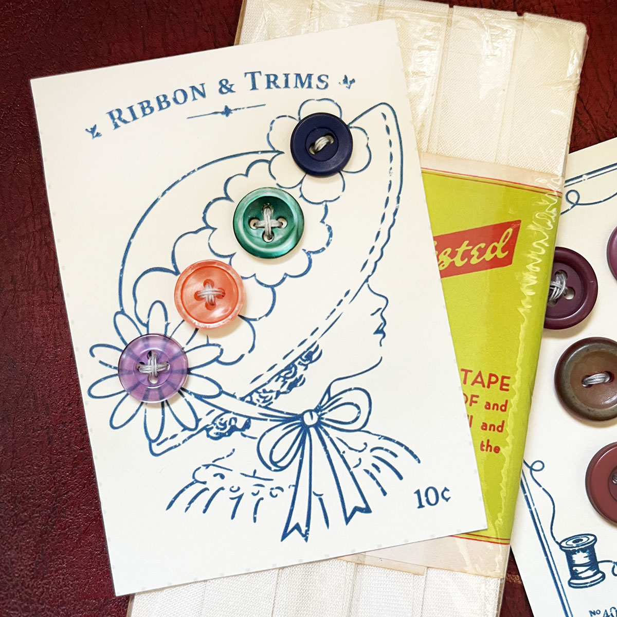 Victorian line art in navy blue of a girl with four flowers on her hat with buttons sewn onto the flower centers in purple, coral, emerald green and navy. Button carda and another card with maroon buttons and a hit of vintage ivory sewing tape in the package shown on a maroon background.
