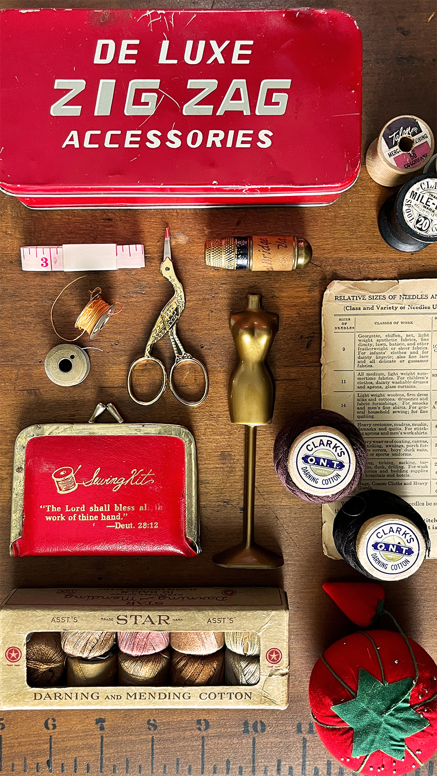 Flatlay overhead shot of vintage notions including a Zig Zag Accessories tin, measuring tape, a vintage thimble with container, thread spools, a STAR darning box kit, tiny purse size sewing kit with a bible verse on it and a tomato pin cushion. Elements flow from red to brass to warn kraft paper.