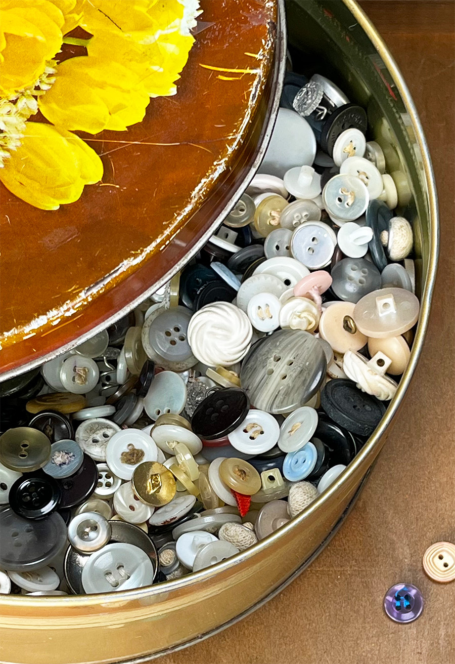 Overhead photo of an open vintage button tin filled with buttons in blues, whites, grays, black, with a peak of the lid with yellow tulips on it.