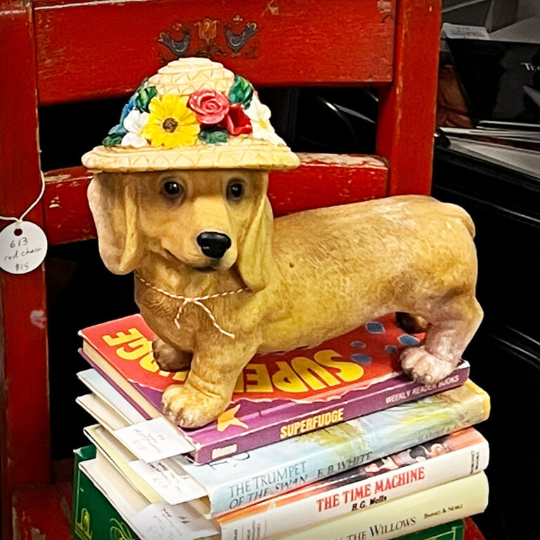 Vintage dog figurine wearing a flowered hat displayed on a stack of books atop a weathered red chair. Editorial Pinterest graphic for a personal essay about closing an antique booth and beginning a new creative chapter.