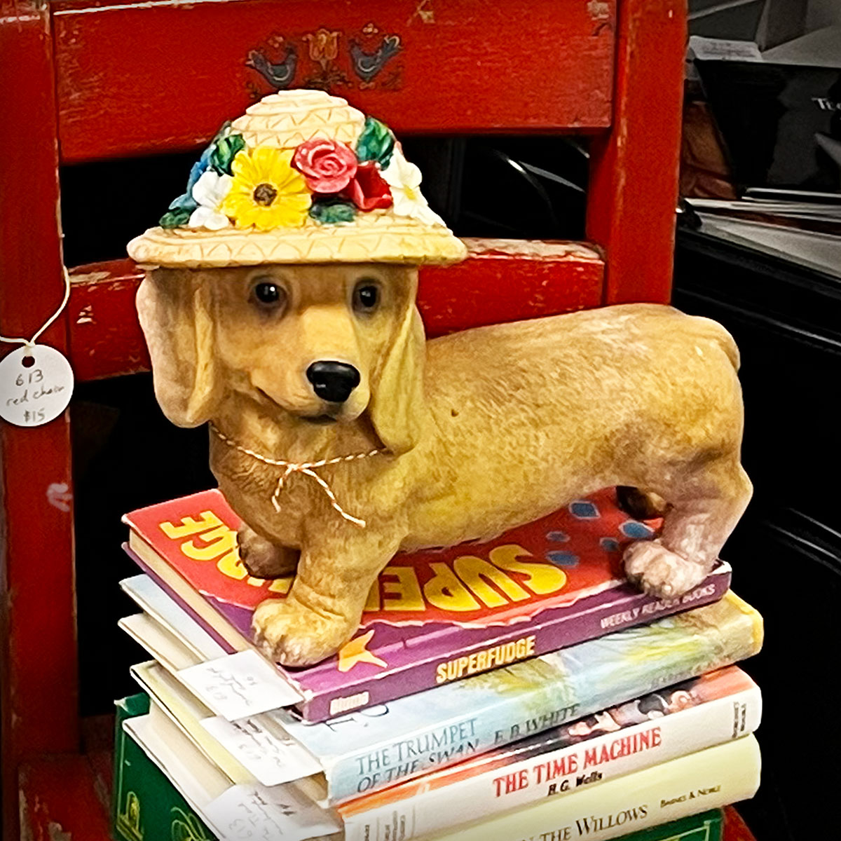 Vintage dog figurine wearing a flowered hat displayed on a stack of books atop a weathered red chair. Editorial Pinterest graphic for a personal essay about closing an antique booth and beginning a new creative chapter.