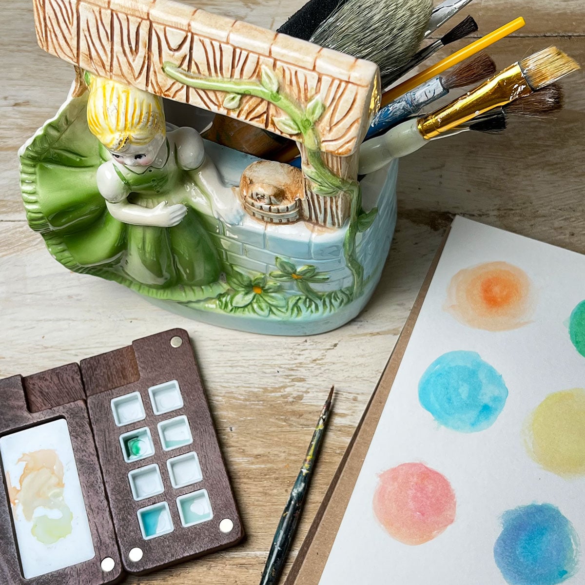 Vintage girl at the well ceramic planter holding paint brushes with a watercolor swatch of circles painted and a small wooden paint mixing palette.