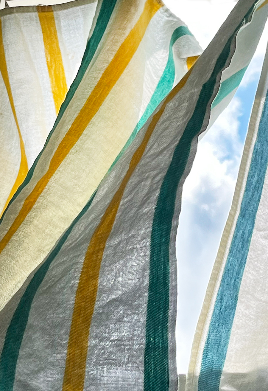 Sunlight shines through vintage white dish towels with faded blue, turquoise, and yellow stripes hanging as simple summer kitchen curtains.