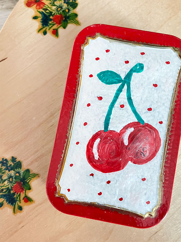 Closed lid of an upcycled altoid tin turned ring holder with hammered finish and two painted red cherries with green stem and one leaf on a white background with red dots using oil based acrylic markers.