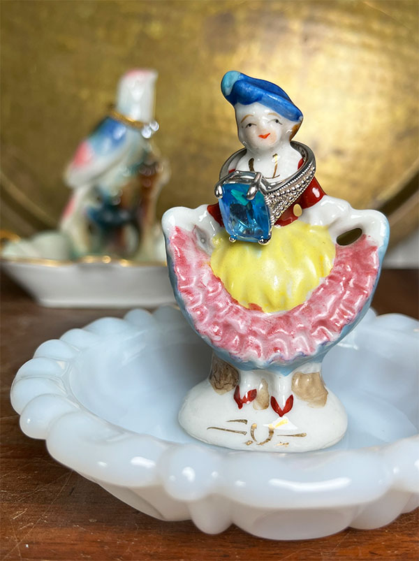 Vintage porcelain girl figurine used as a ring holder on a white milk glass butter pat dish.