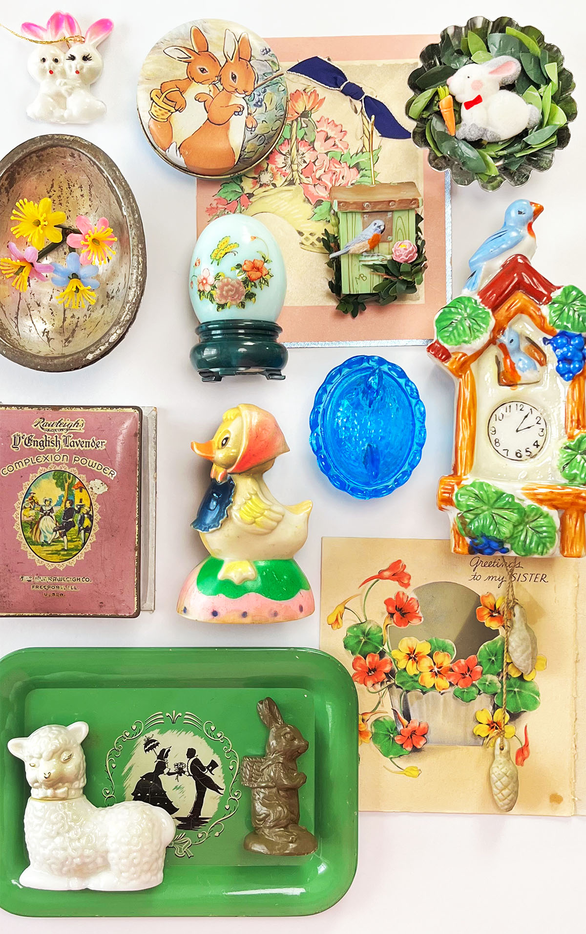 Vintage Easter decor flat lay with small pastel objects arranged in a spring vignette, including a white Avon lamb bottle, pull toy chick, chocolate egg mold, blue glass dish, and handmade rabbit tin magnet in soft blues, pinks, seafoam green, and warm brown tones.