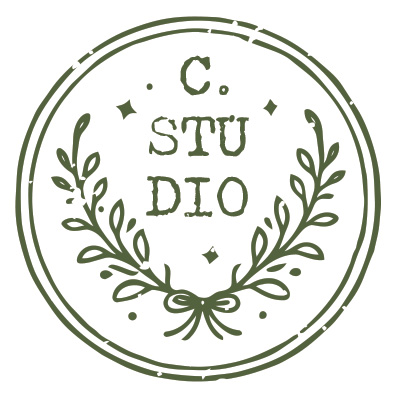 Circular botanical maker's mark featuring a leafy wreath tied with a bow, distressed double-ring border, and stacked "C. STUDIO" lettering in the center, illustrated in muted moss green.