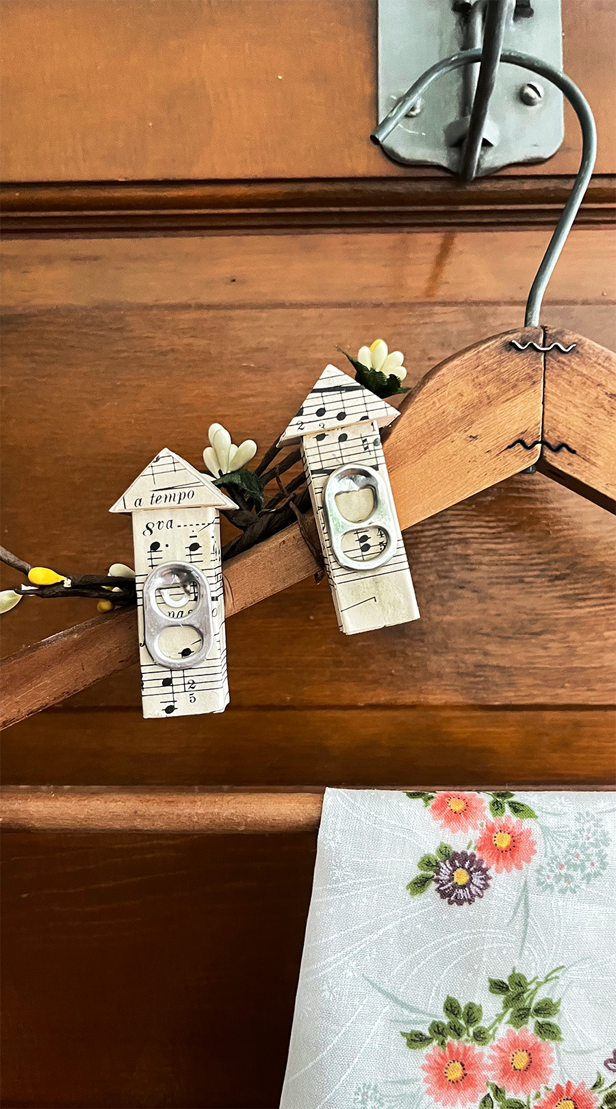 Old wooden hanger with two miniature birdhouses attached to the upper left, covered in vintage sheet music with soda can pull tabs as the birdhouse openings. A vintage peach and deep purple handkerchief hangs over the center of the wooden hanger.