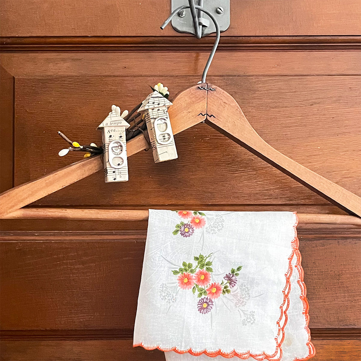 Old wooden hanger with two miniature birdhouses attached to the upper left, covered in vintage sheet music with soda can pull tabs as the birdhouse openings. A vintage peach and deep purple handkerchief hangs over the center of the wooden hanger.