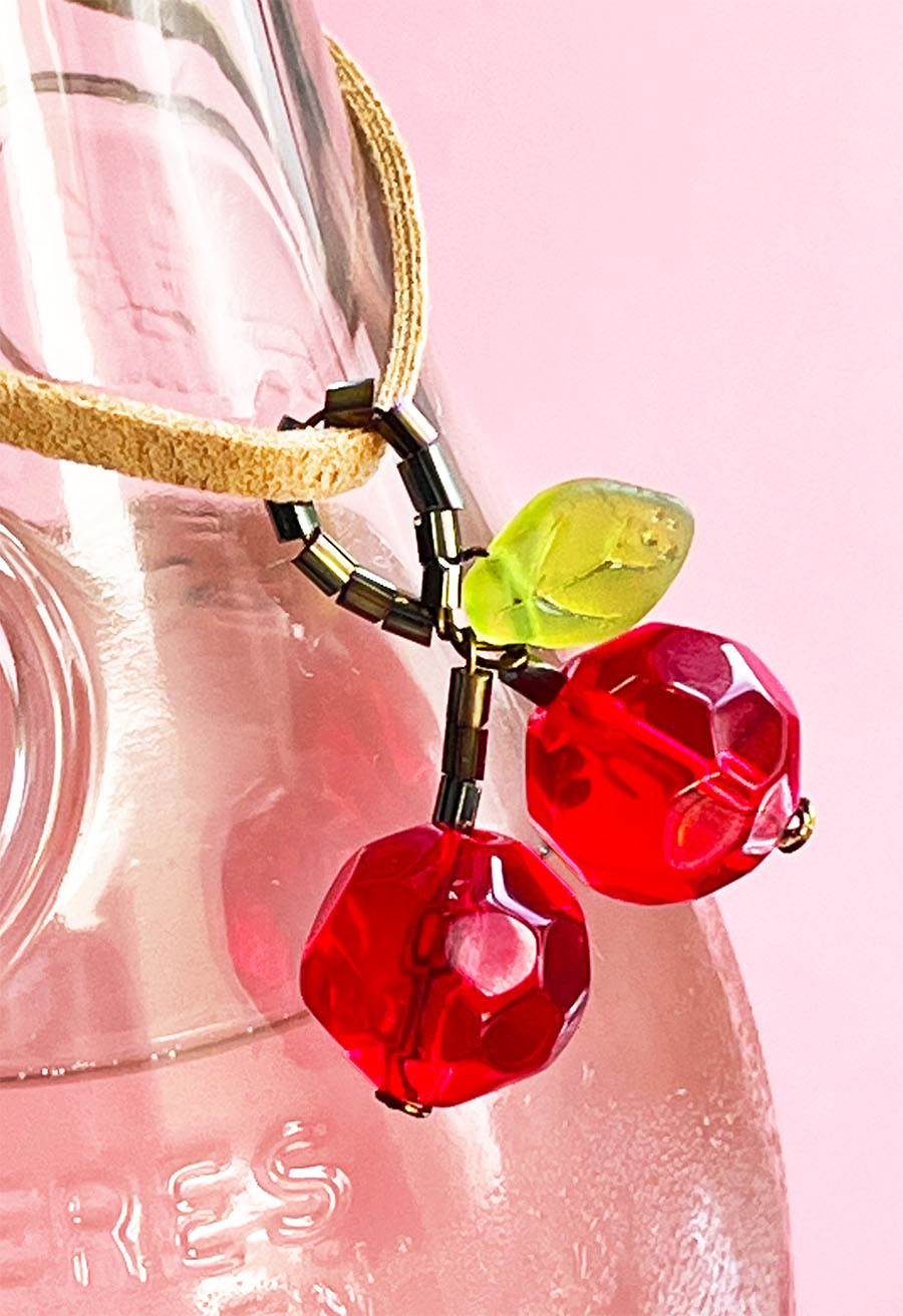 Handmade beaded cherry charm with faceted red beads and a green leaf, hanging from a suede cord on a pink lemonade bottle.
