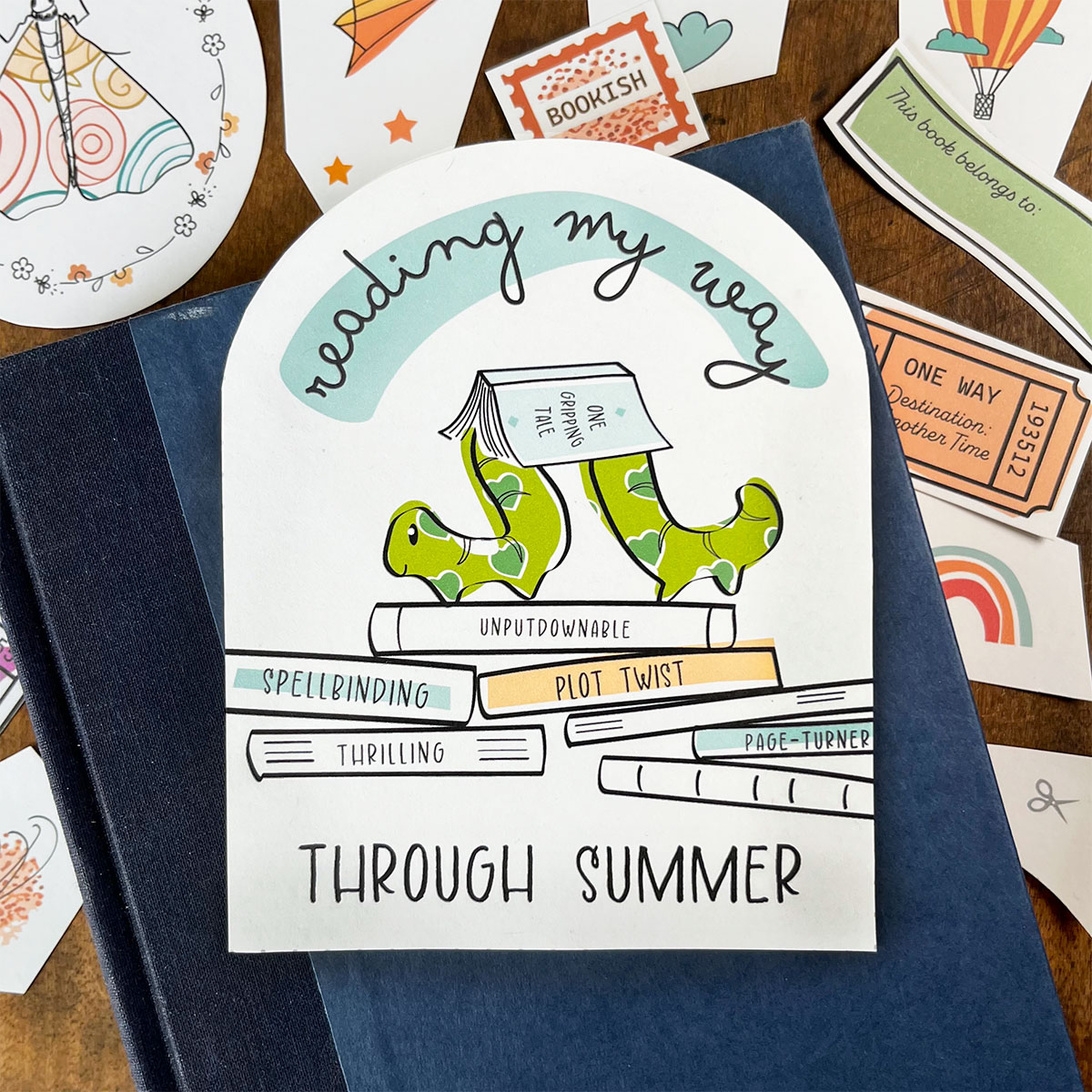 Reading my way through summer featured on a blue book cover. It has an inch worm carrying an open book on it's back and around it are other ephemera and reading paper buts that easily fit into the reading tote made in week 1, Summer 2026 installment of The Paper Snail Club.