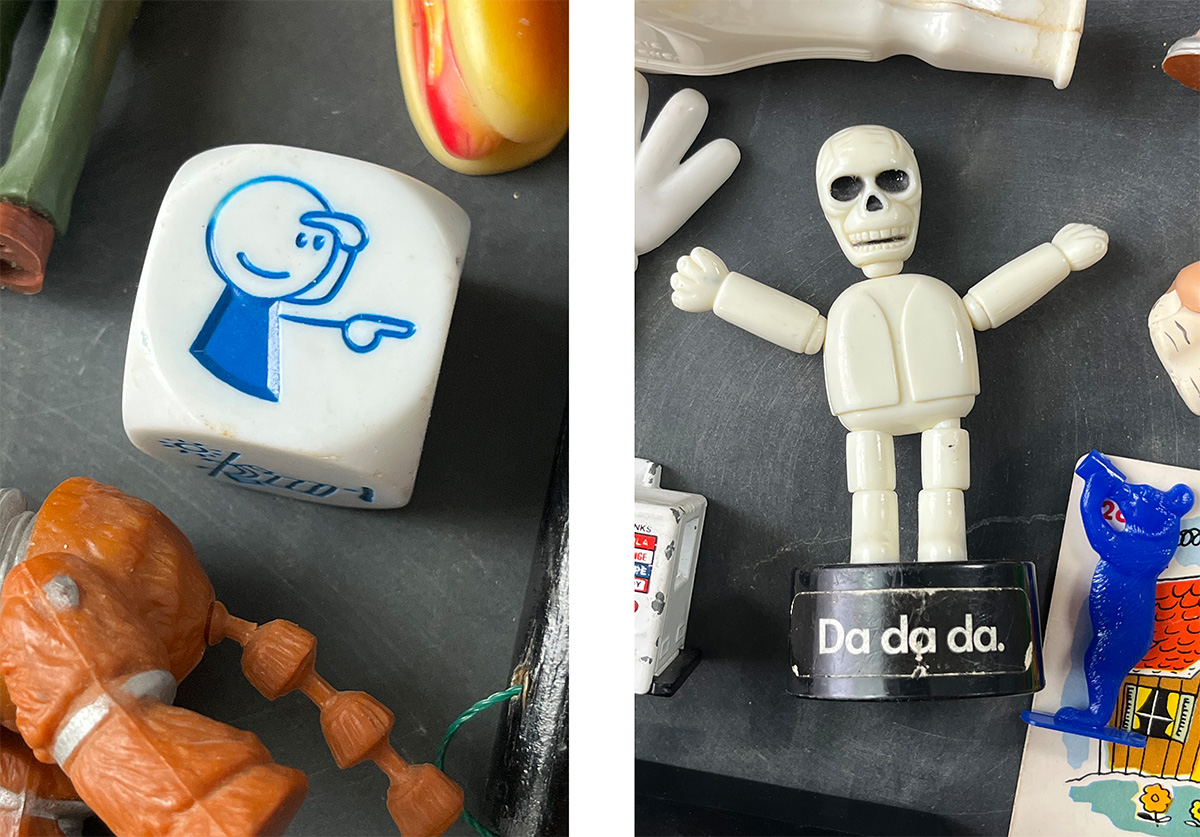 Photo 1: Vintage game die featuring a blue pointing figure surrounded by small nostalgic plastic toys, including an orange jointed figure and colorful game pieces arranged on a dark surface. Photo 2: Vintage articulated skeleton toy standing on a black "Da de da." display base, surrounded by small novelty figures and collected curiosities arranged on a dark tabletop.