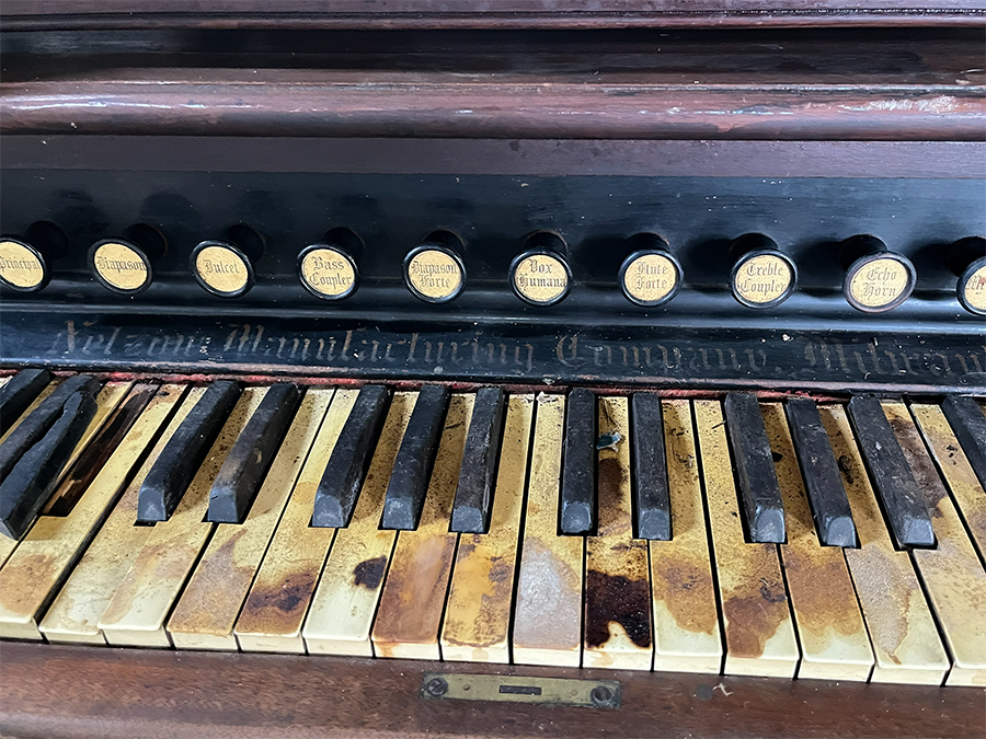 A horizontal shot of a worn, aged and keys from an old organ in disrepair at a thrift store. It has black vintage round pull knobs above the keys that give sound options like Base Coupler and Echo Horn. The writing on the round knobs is in faded white caligraphy as well as the name of the organ manufacturing company.