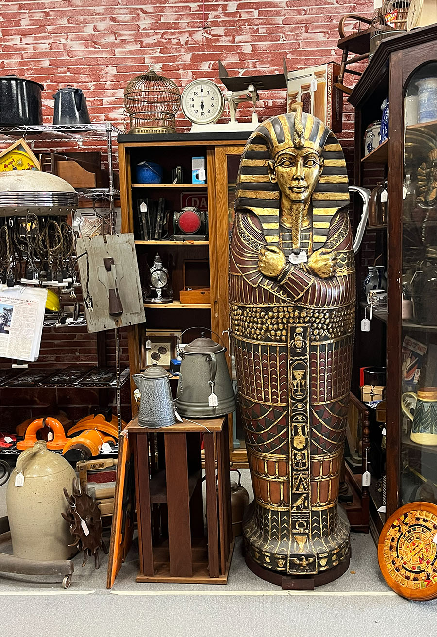 An Egyptian sarcophagus standing in an antique store booth. The booth is about 10 feet wide and filled with cabinets, antiques and curiosities like enamelware kettles, large stone jugs, crates, a vintage meat scale, deorative birdhouse and chair perched on the hightest shelf top and a cabinet filled with collectable vintage ale mugs and glass pieces.