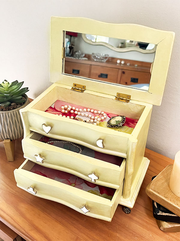 Vintage jewelry box refinished in soft champagne with an open mirrored lid, floral-lined drawers, and handmade white clay charm pulls edged in gold, including hearts, stars, a moon, clover, spade, and diamond.