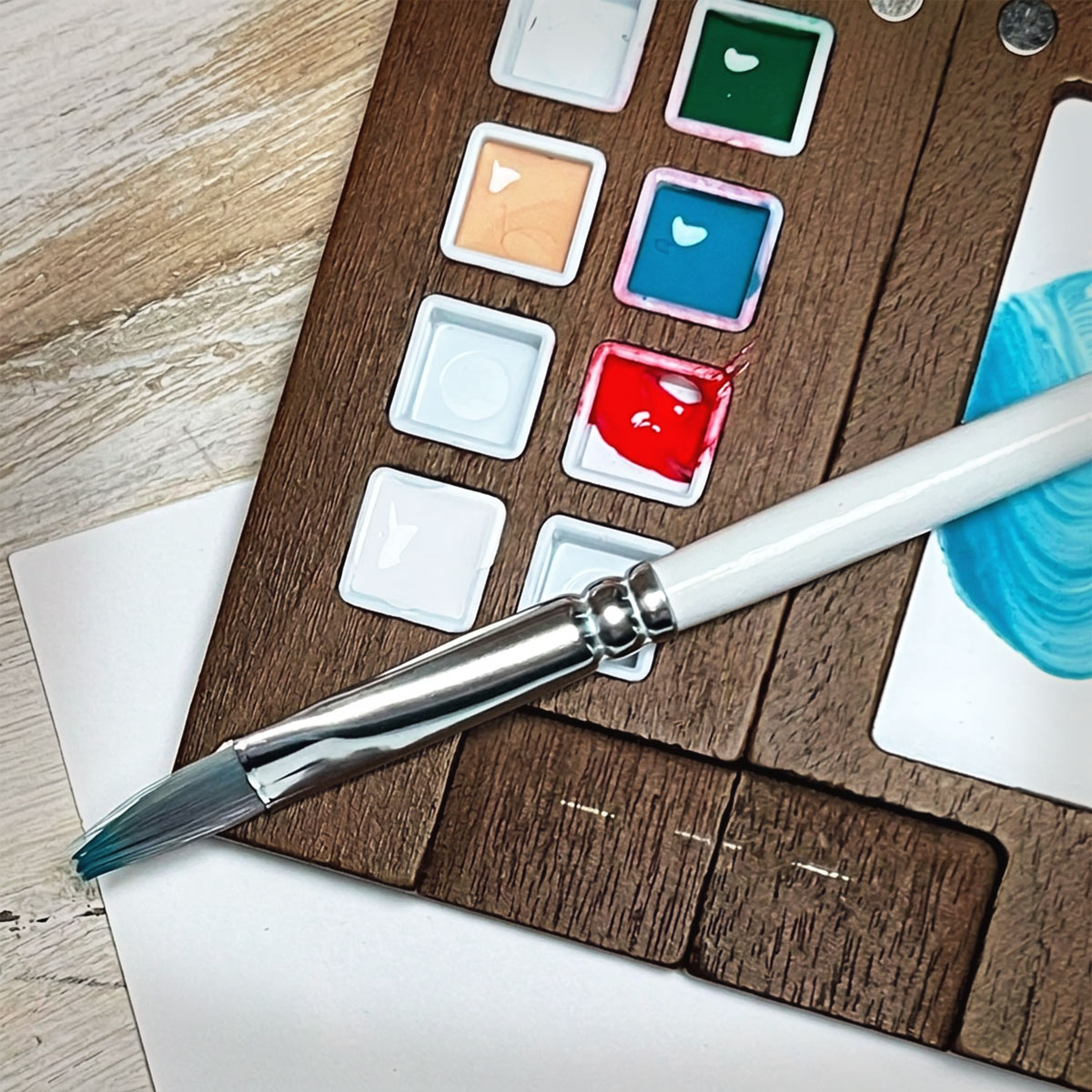 Closeup of a miniature wooden paint palette with a paint brush and paint in the wells of aqua, white, beige and red.