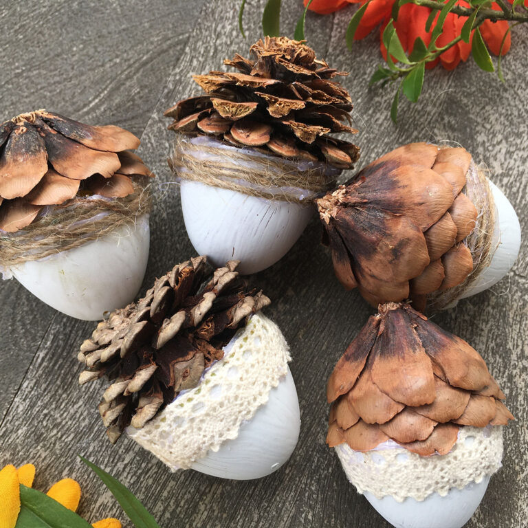 Five crafted acorns on a dark background with hints of fall orange and yellow florals to the side. The acorns are made from small plastic Easter eggs painted with white chalkpaint and topped with half of a pine cone.