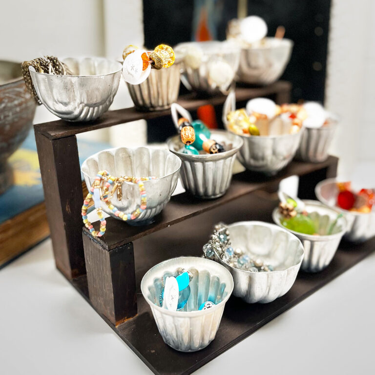 Square right facing feature image of a DIY three tiered organizer made with scrap wood and 12 vintage round and scalloped tart tins facing to the left. There are four on each tier and they hold beads, small jewelry and bracelets with tags for sale in a vintage antique booth.