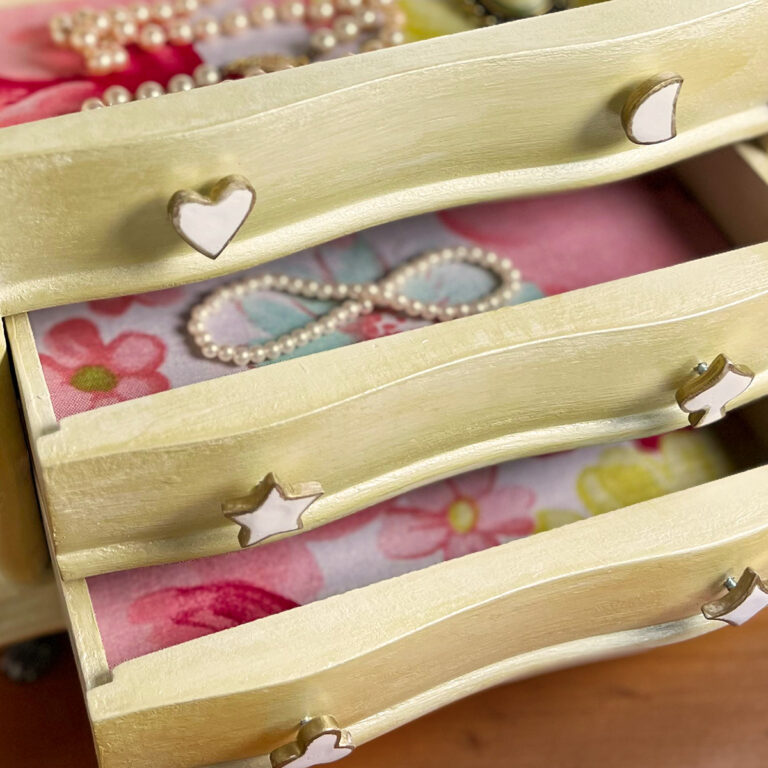 Close-up of a thrift store jewelry box makeover with a soft champagne painted finish, floral fabric-lined drawers, and handmade white clay charm pulls edged in gold.