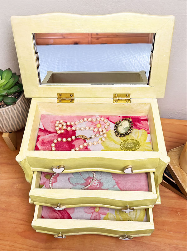 Slightly overhead shot of a vintage jewelry box refinished in soft champagne with an open mirrored lid, floral-lined drawers, and handmade white clay charm pulls edged in gold, including hearts, stars, a moon, clover, spade, and diamond.