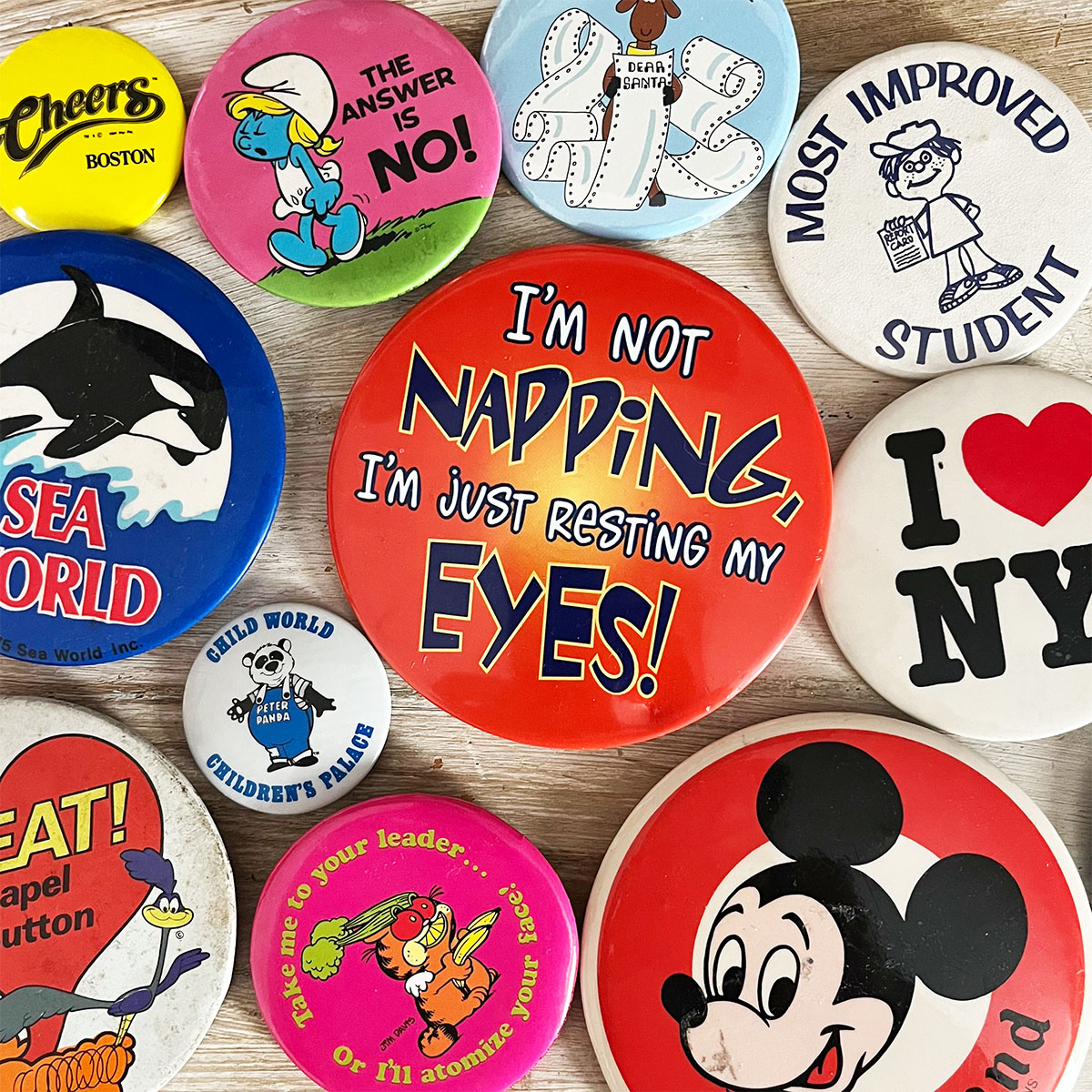Vintage pin-back buttons arranged in a colorful flat lay, featuring retro slogans, characters, travel souvenirs and childhood keepsakes, including Sea World, I Love NY, Mickey Mouse, Smurf and Road Runner buttons.