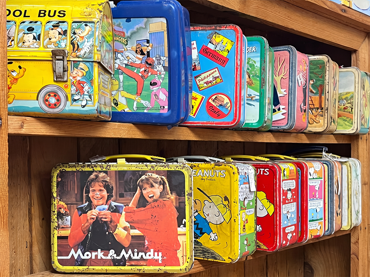 Metal vintage kids lunch boxes lining two antique store shelves. In the front is a school bus Mickey Mouse lunch box and Mork & Mindy lunch box followed by Peanuts and other.