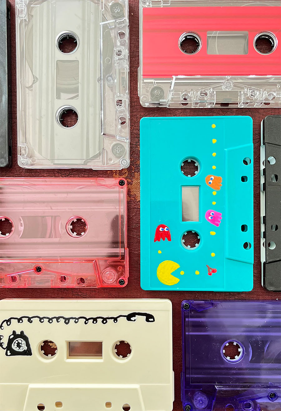 Colorful vintage cassette tapes arranged on a dark red background, including an aqua tape hand-painted with Pac-Man, ghosts, dots and cherries, and an ivory tape with a hand-drawn rotary telephone and curly cord.