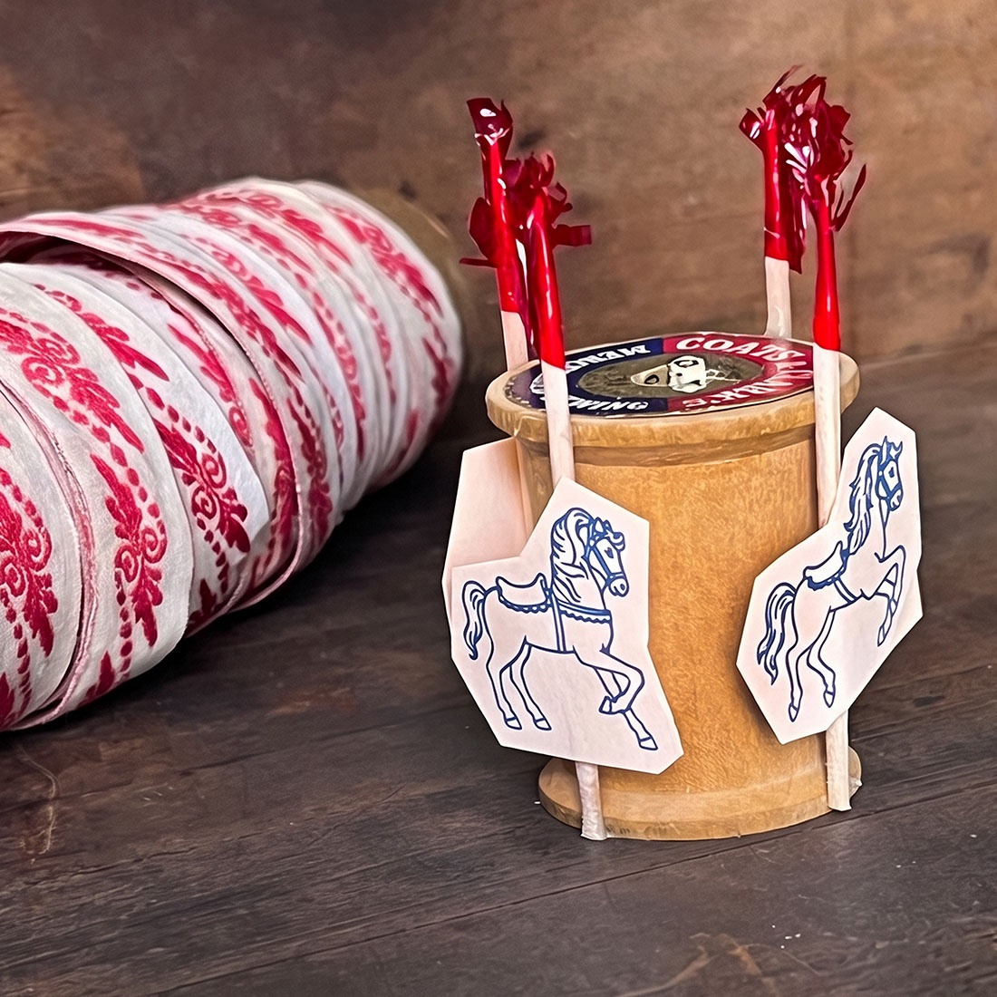Vintage wooden thread spool transformed into a miniature carousel with blue illustrated paper horses on decorative picks, styled beside red-and-ivory vintage ribbon.