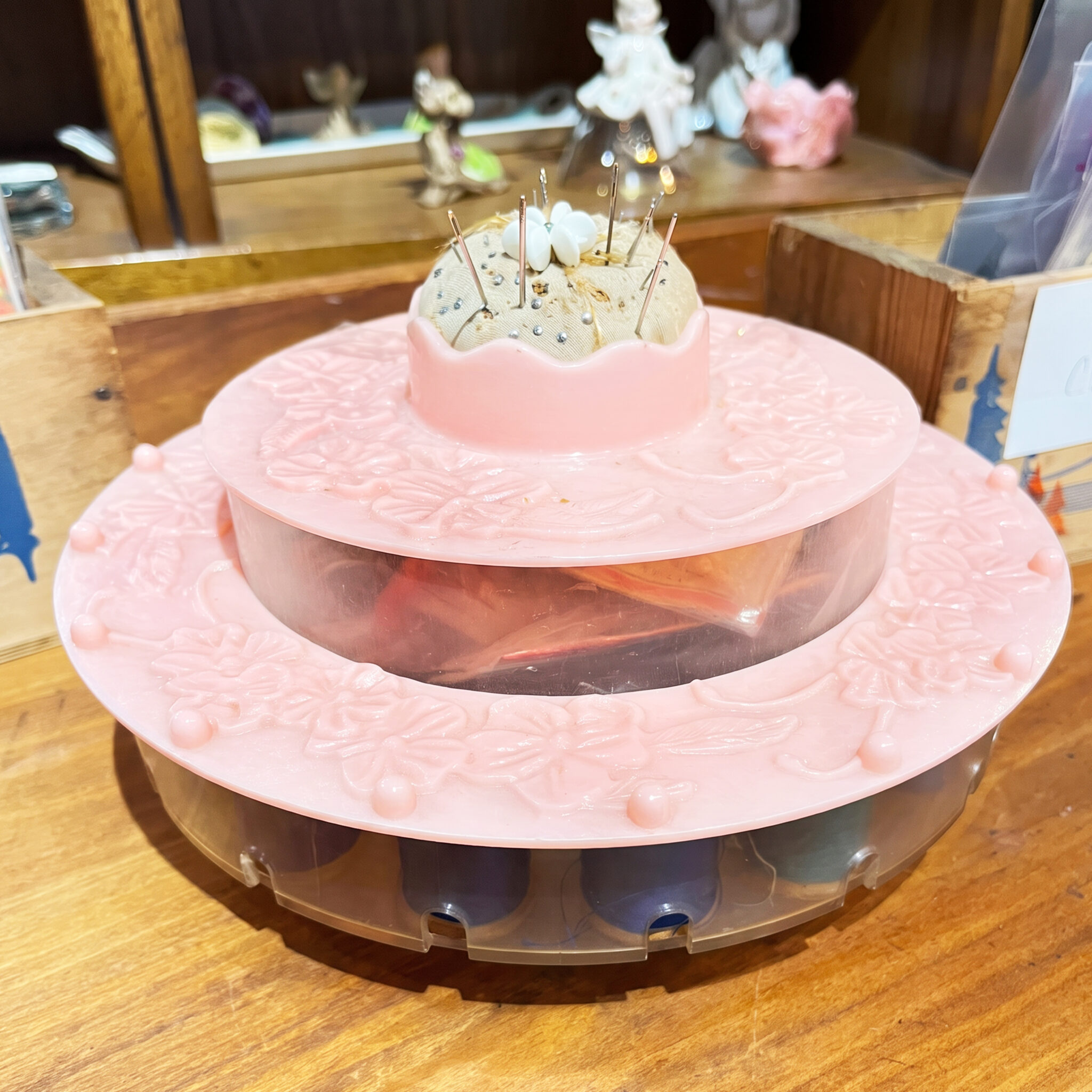Vintage pink tiered sewing notion holder with translucent compartments for thread and a built-in pincushion filled with pins.