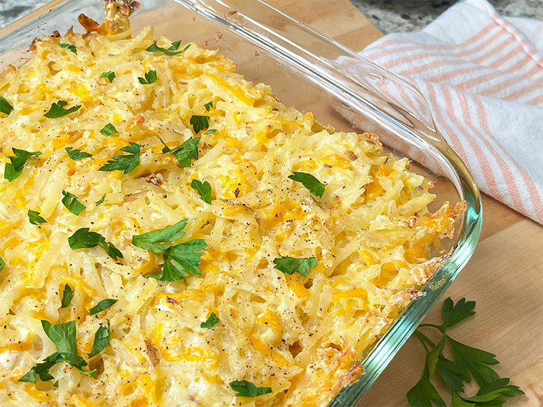Closeup of a 13" x 9" casserole of Baked Cheesy Hash Brown Potatoes with browned cheesy top layer dotted with parsley on a wood serving board.