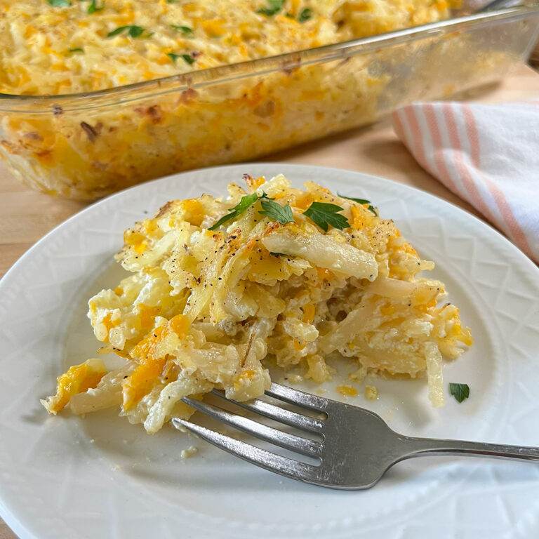 Closeup of a white dish with fork of Baked Cheesy Hash Brown Potatoes with the full casserole in the background.