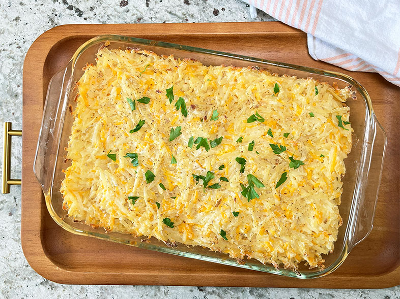 Fresh from the oven cooling on a wood serving board, Baked Cheesy Hash Brown Potatoes, golden brown dotted with parsley garnish.