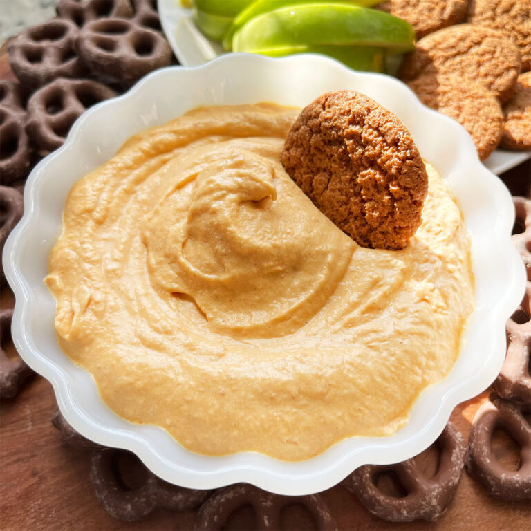 A fluted bowl with pumpkin fluff dip with dippers like chocolate covered pretzels, slices of green apple and gingersnaps.