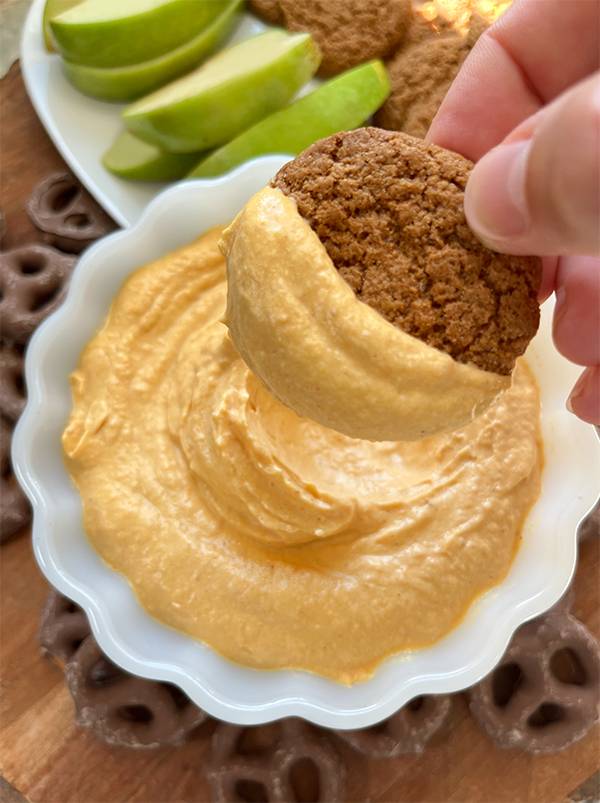 A fluted bowl with pumpkin fluff dip with dippers like chocolate covered pretzels, slices of green apple and gingersnaps. Dipping a gingersnap into the bowl.