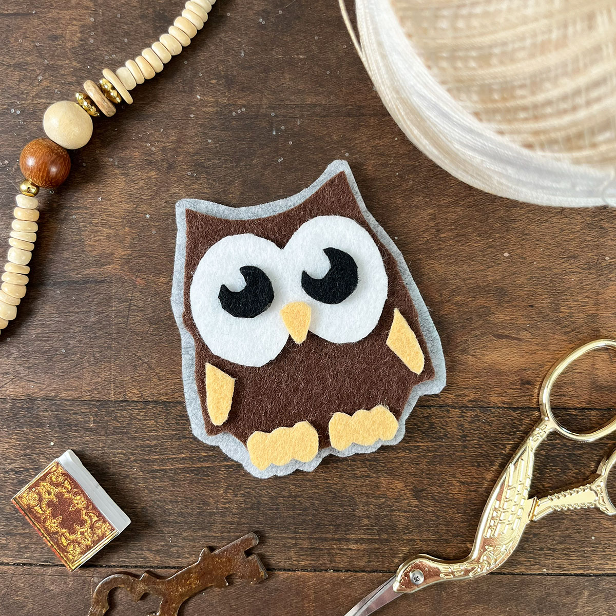 Handmade owl feltie (felt applique) on a warm wood surface with a natural beaded necklace, a tiny book, a vintage key and embroidery scissors nearby.